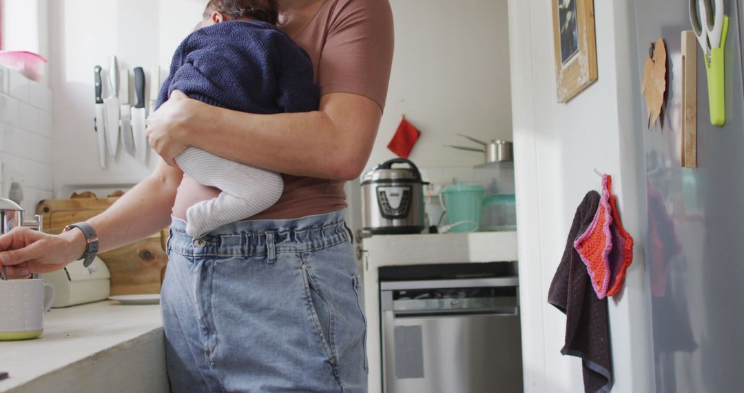 Parent holding baby in kitchen performing daily chores - Free Images, Stock Photos and Pictures on Pikwizard.com