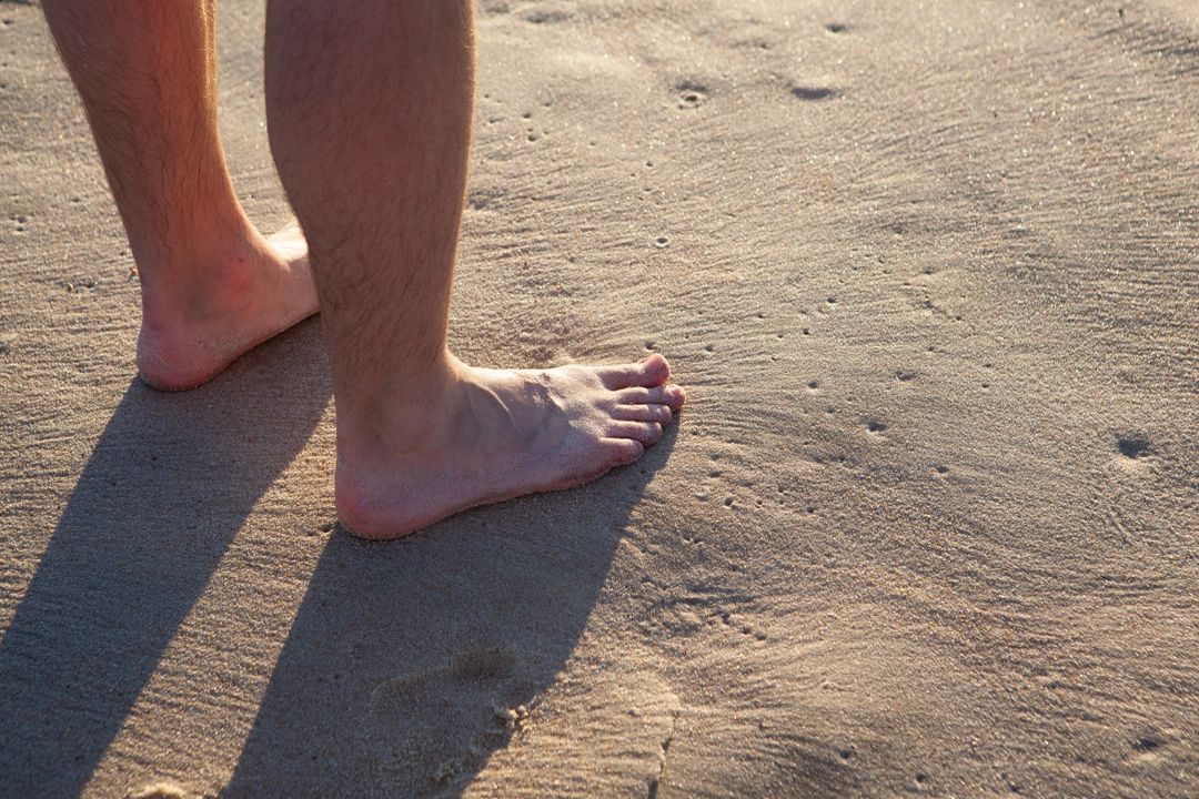 Standing barefoot on sunlit beach sand, close-up of lower legs and toes casting shadow