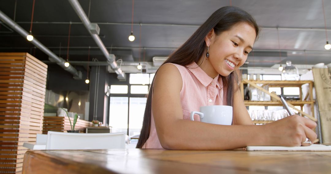 Smiling Woman Writing at a Cafe Holding Coffee Mug - Free Images, Stock Photos and Pictures on Pikwizard.com