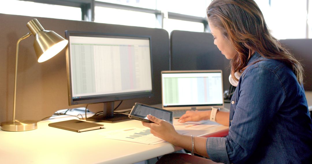 Woman working at desk reviewing analytics and spreadsheets on dual monitors with tablet