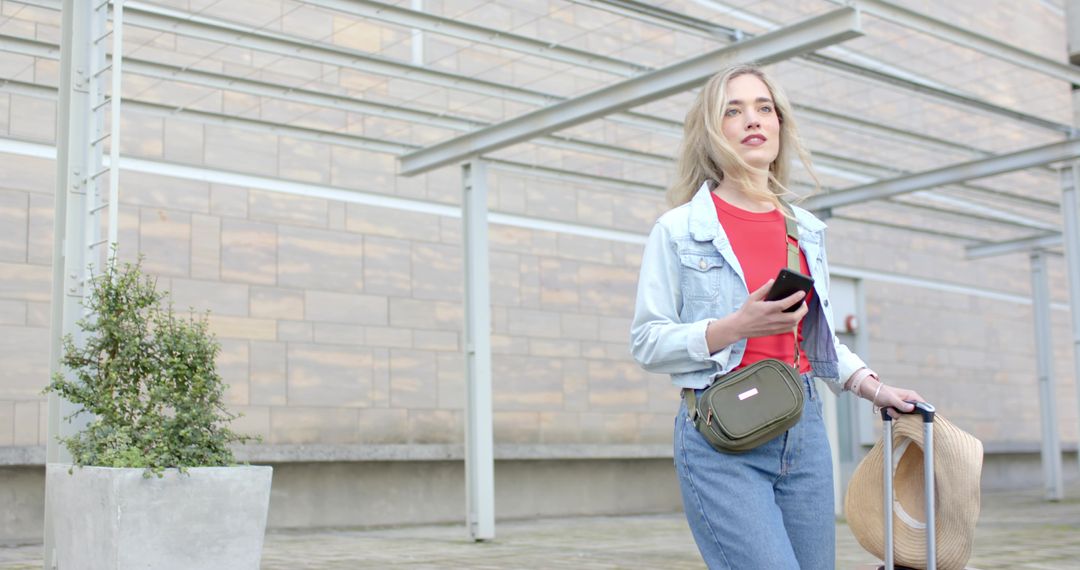 Young woman traveling with rolling suitcase and straw hat using smartphone in urban plaza