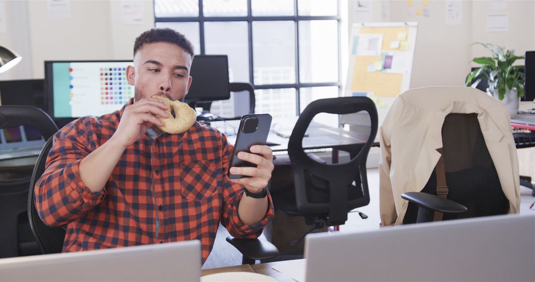 Young Professional Eating Bagel While Using Smartphone in Modern Office - Free Images, Stock Photos and Pictures on Pikwizard.com