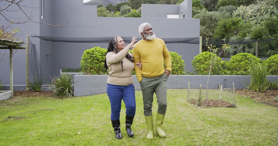 Senior couple walking arm-in-arm across backyard lawn pointing toward garden in boots