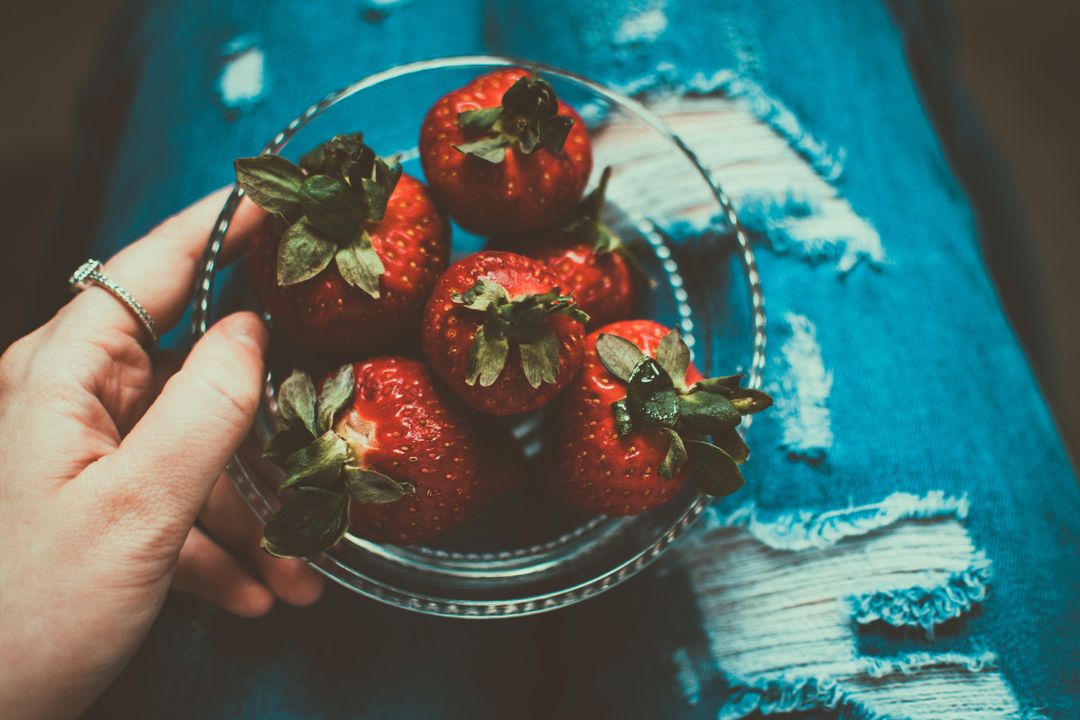 Hand holding bowl of ripe strawberries on ripped denim lap with ring, moody rustic snack