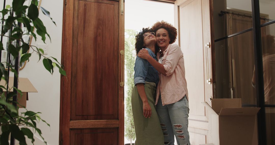 Women embracing while entering new home with moving boxes in sunlit foyer