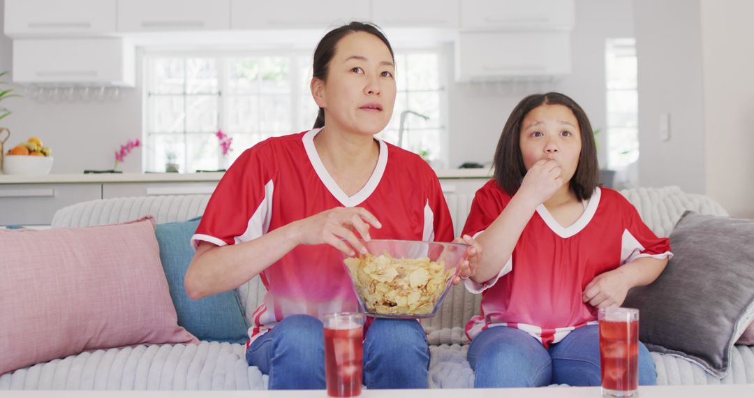 Image of happy asian mother and daughter eating chips and watching match in tv - Free Images, Stock Photos and Pictures on Pikwizard.com