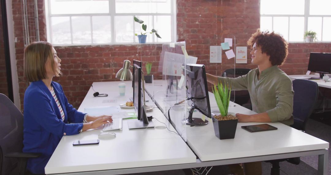 Asian Woman Typing While Colleague Placing Sticky Note Across Clear Office Divider in Loft