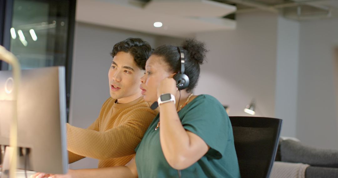 Multicultural coworkers collaborating at desktop, pointing at screen and adjusting headset