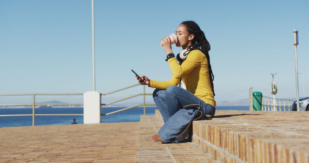 Young Woman Enjoying Coffee by the Sea Using Smartphone - Free Images, Stock Photos and Pictures on Pikwizard.com