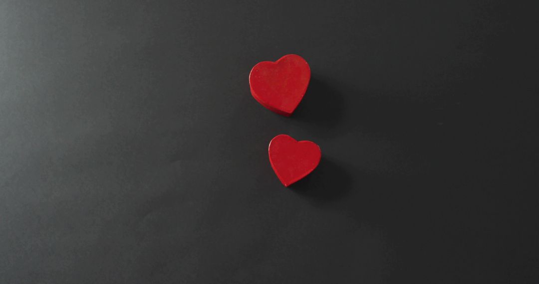 Two red heart blocks resting on dark matte surface casting shadows, minimal composition