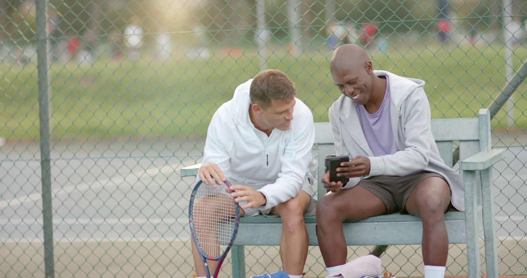 Two Men Sharing a Mobile Phone on a Tennis Court Bench - Free Images, Stock Photos and Pictures on Pikwizard.com