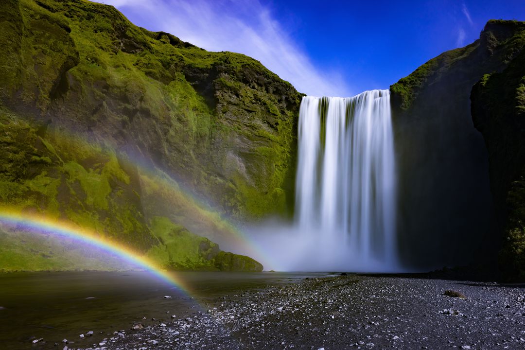 Cascading waterfall with double rainbow over moss-covered cliffs and black pebble shore