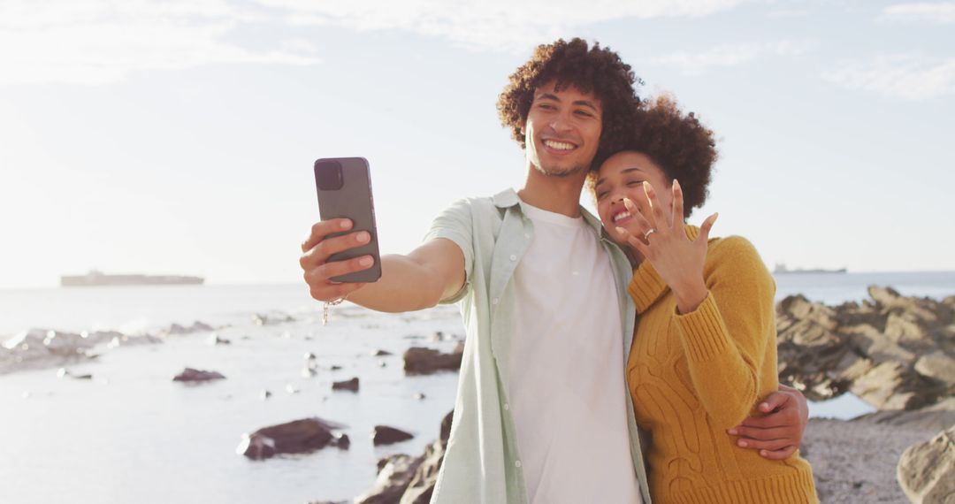 African american couple taking a selfie and showing their ring standing on the rocks near the sea - Free Images, Stock Photos and Pictures on Pikwizard.com