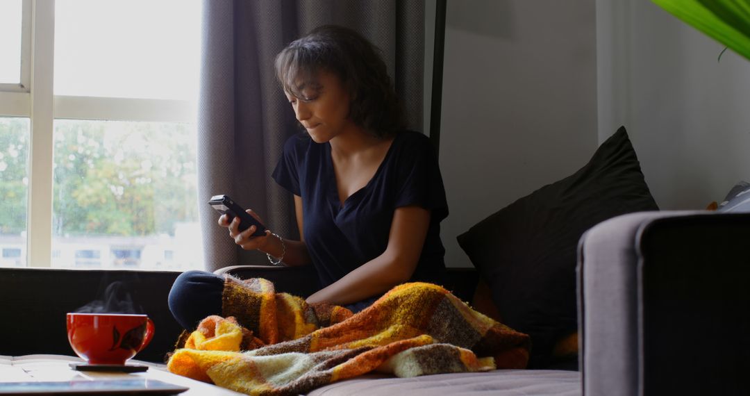 Woman with Blanket Browsing Smartphone by Window in Cozy Apartment - Free Images, Stock Photos and Pictures on Pikwizard.com