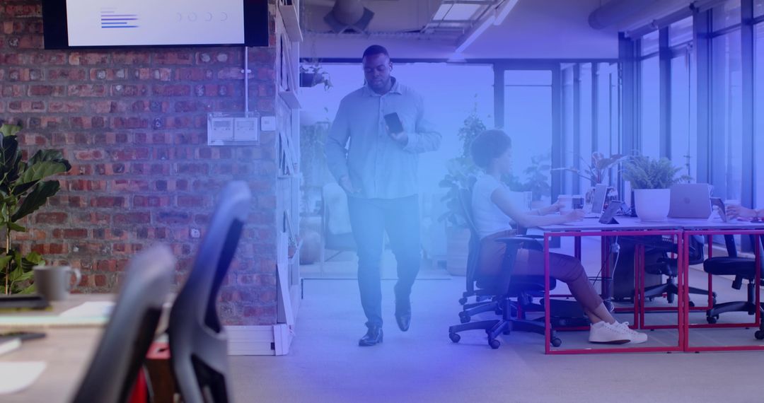 Colleague walking through modern open-plan office checking smartphone near red workstations