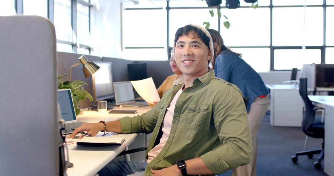 Young professional smiling in open-plan office with diverse team collaborating on paperwork