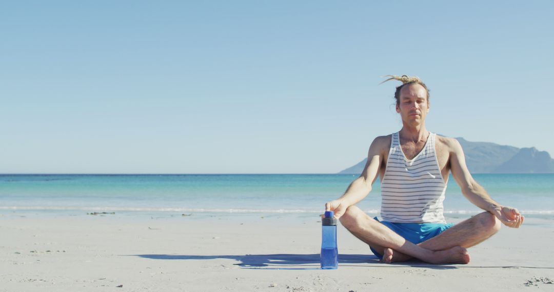 Man meditating on beach with water bottle under clear sky - Free Images, Stock Photos and Pictures on Pikwizard.com