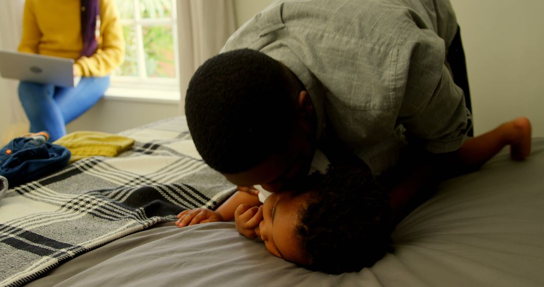 Father Kissing Child on Bed with Woman Using Laptop in Background - Free Images, Stock Photos and Pictures on Pikwizard.com