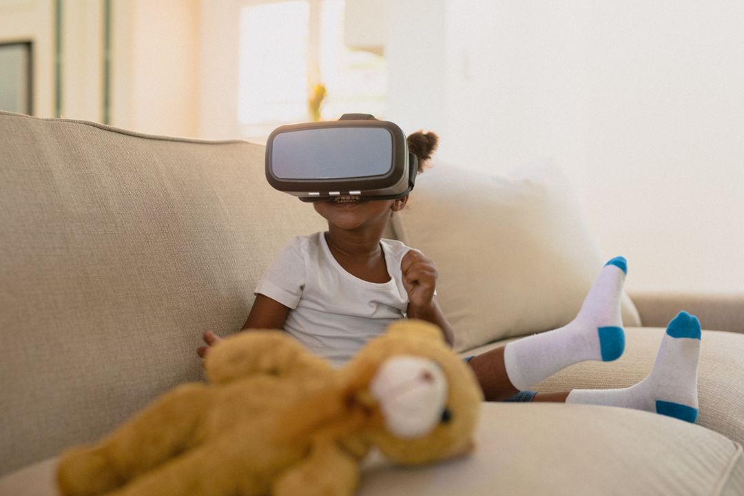 African american girl wearing vr headset and sitting on sofa with teddy bear - Free Images, Stock Photos and Pictures on Pikwizard.com