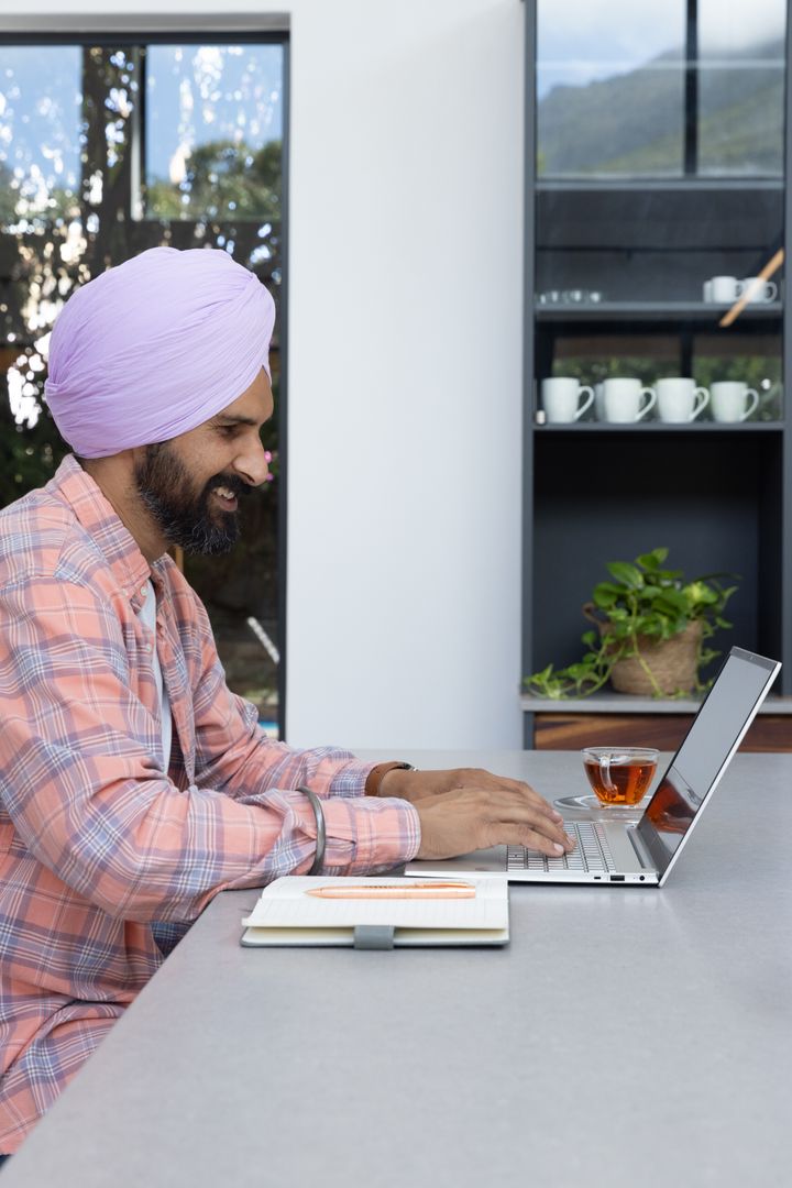Professional Man Typing on Laptop in Modern Kitchen with Tea and Notebook