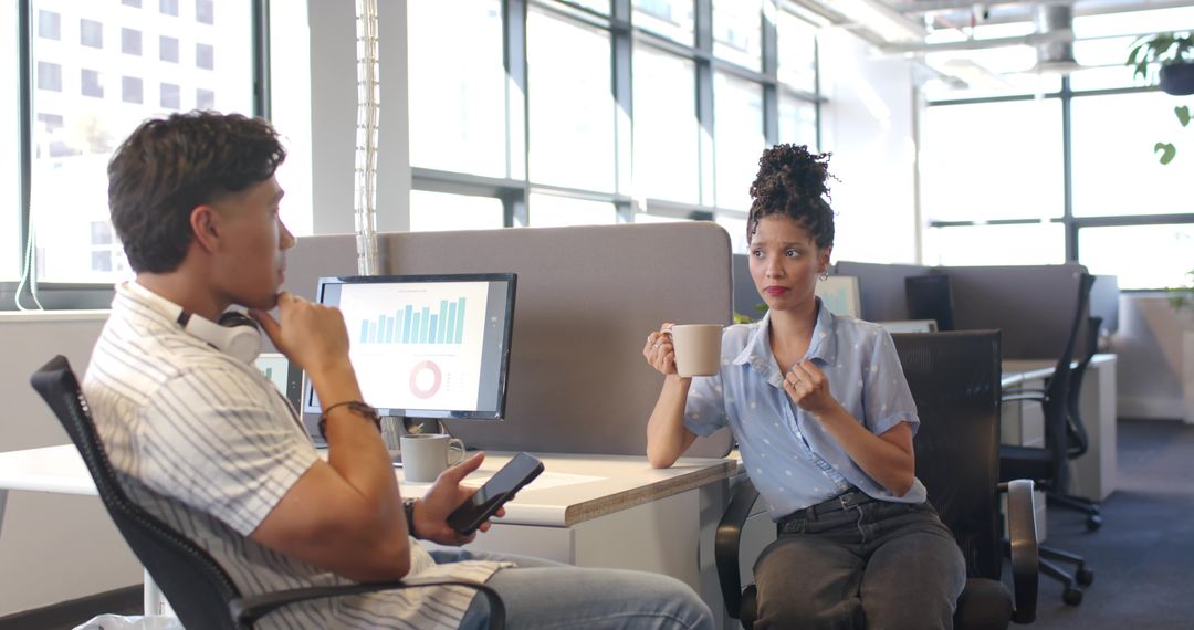 Diverse coworkers discussing charts over coffee in sunlit open-plan office workspace
