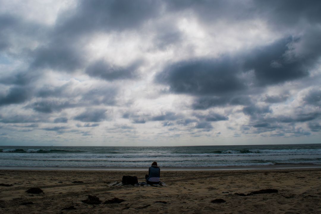 Solitary Figure Sitting on Cloudy Beach - Free Images, Stock Photos and Pictures on Pikwizard.com