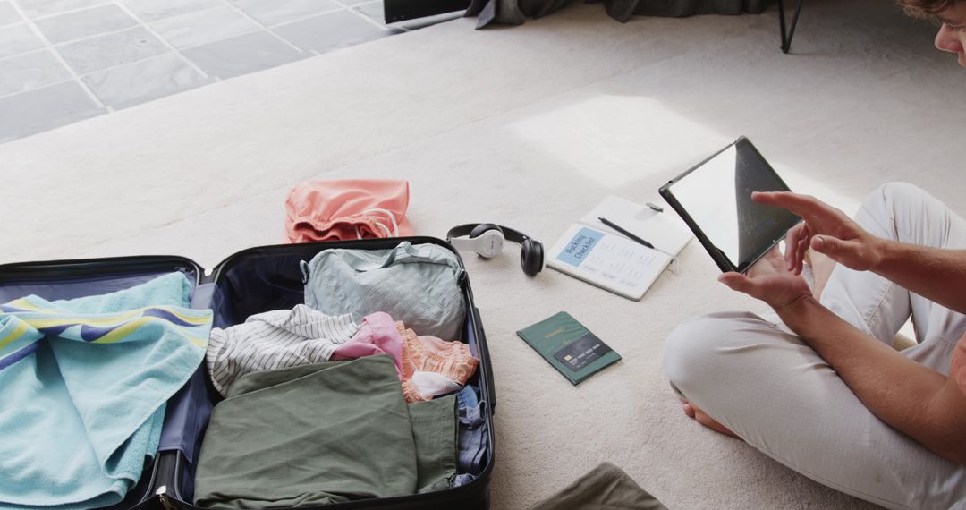 Young Man Packing Carry-On While Using Tablet and Passport for Trip Planning
