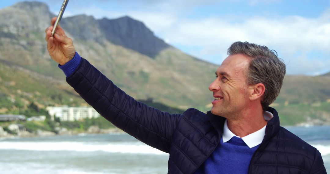 Smiling Middle-Aged Man Taking Selfie at Scenic Beach with Mountains in Background - Free Images, Stock Photos and Pictures on Pikwizard.com