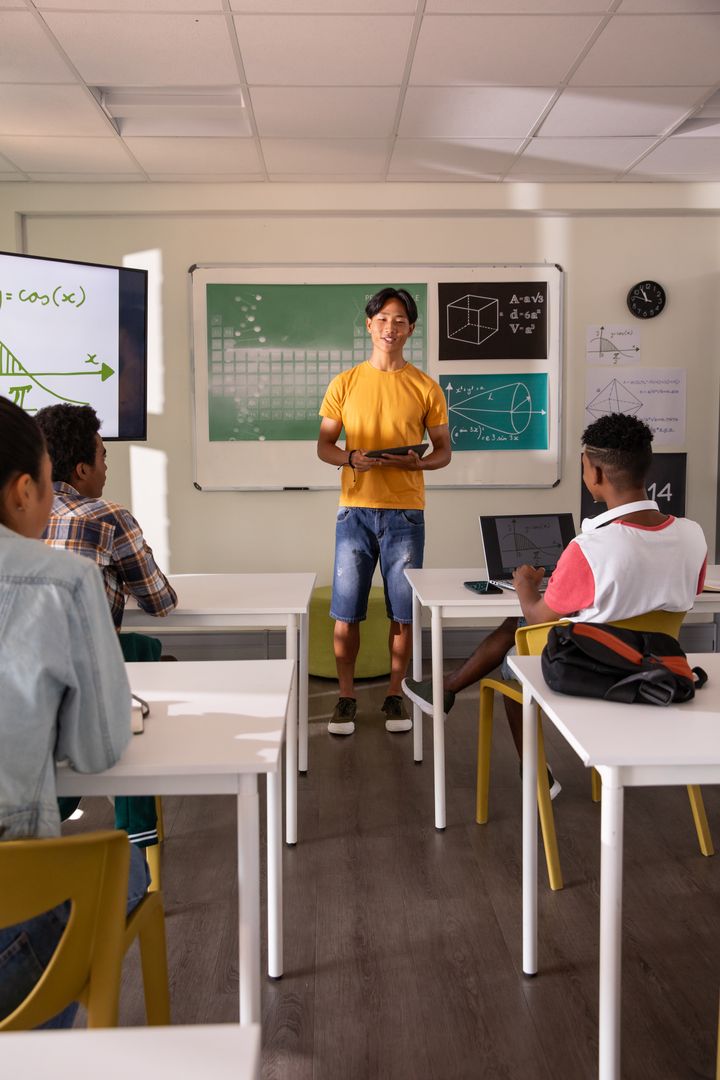 Teenage Asian boy presenting STEM lesson holding tablet in classroom with classmates