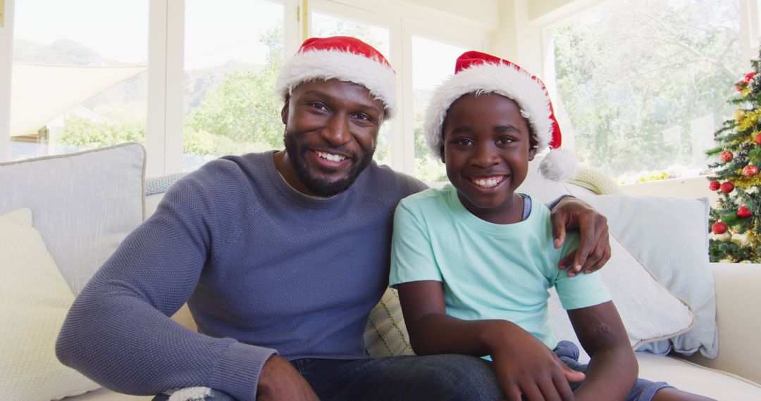 Father and son sitting on couch wearing Santa hats smiling - Free Images, Stock Photos and Pictures on Pikwizard.com