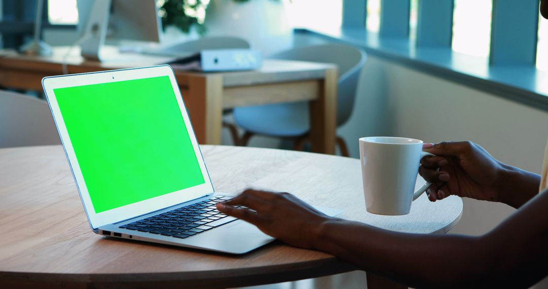 Person Working on Laptop with Green Screen and Holding Coffee in Modern Office - Free Images, Stock Photos and Pictures on Pikwizard.com