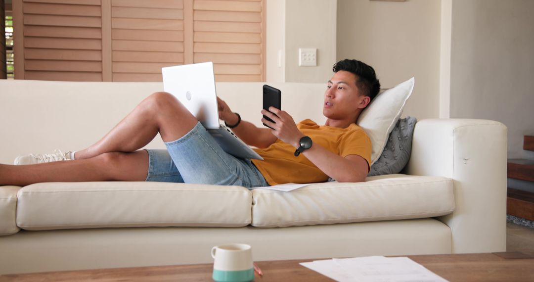 Man Relaxing on Sofa Multitasking with Laptop and Smartphone