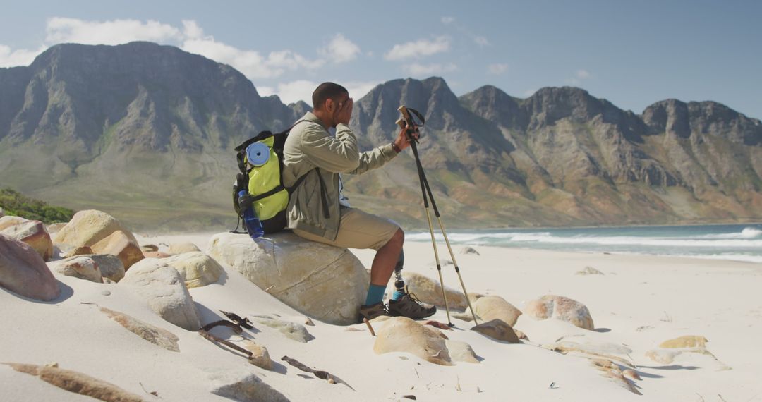 Biracial man with prosthetic leg trekking with backpack and walking poles taking a break on a beach - Free Images, Stock Photos and Pictures on Pikwizard.com