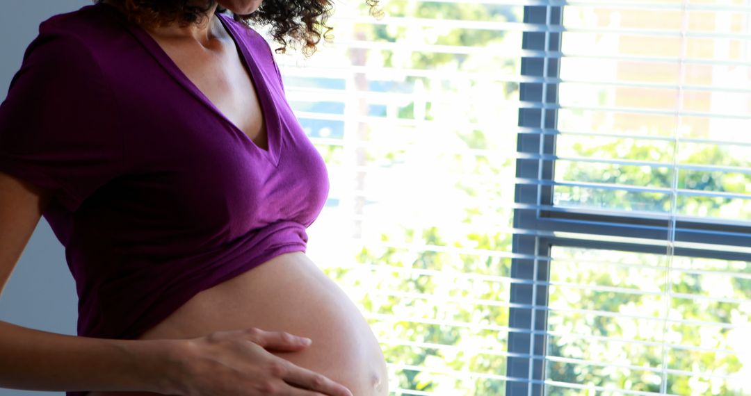 A pregnant woman in purple tenderly holds her belly by a window, embodying maternal anticipation. - Free Images, Stock Photos and Pictures on Pikwizard.com