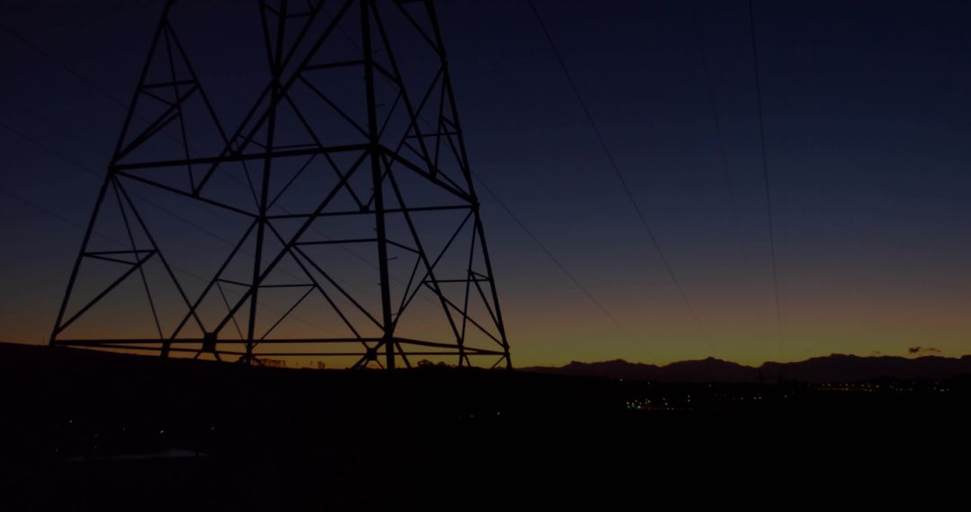 Looming Transmission Tower Silhouette at Dusk Sending Power Lines Across Mountain Horizon