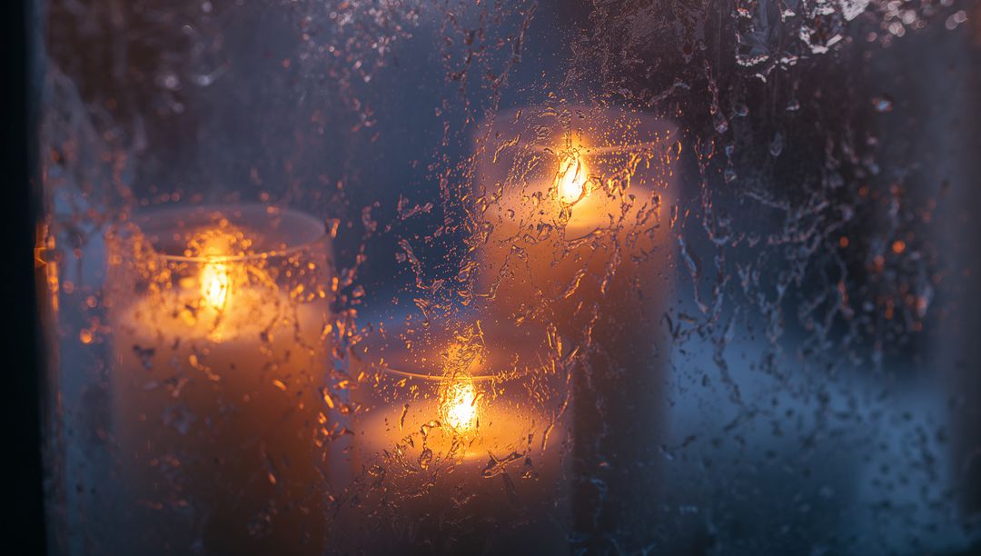 Warm pillar candle glow behind rain-speckled glass creating cozy moody evening ambience