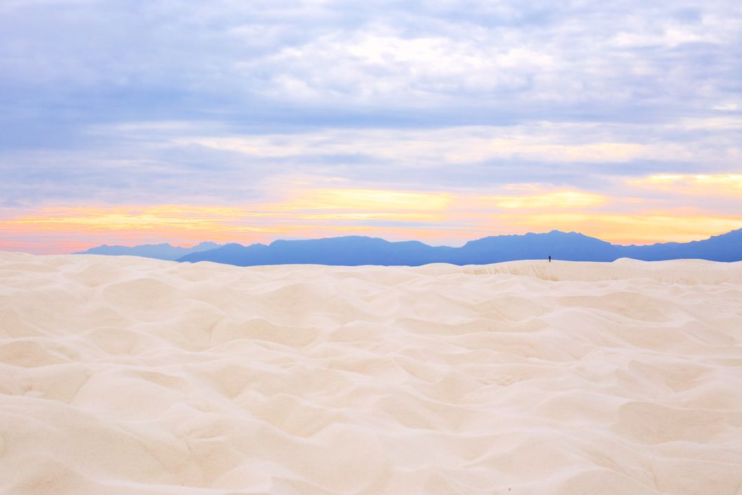 Wandering figure on white sand dunes at pastel sunset with distant mountain silhouette