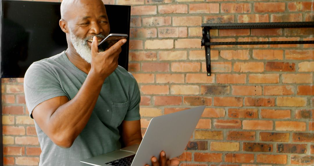 Senior Man Using Voice Command on Smartphone While Holding Laptop in Modern Loft - Free Images, Stock Photos and Pictures on Pikwizard.com