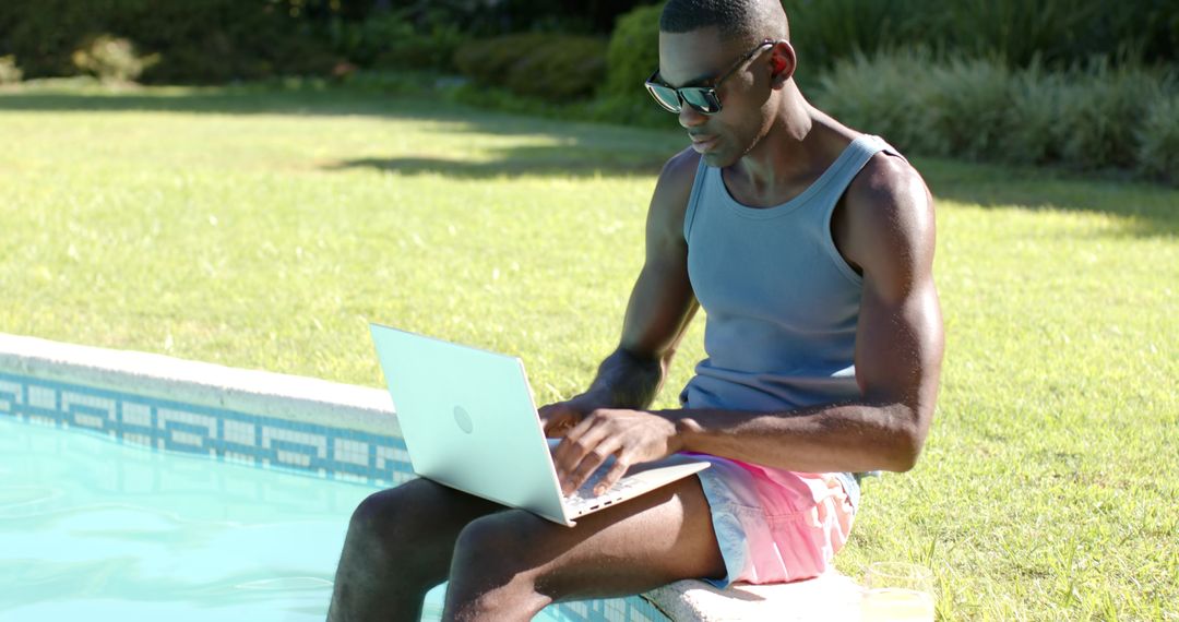 Young African American Man Working Remotely on Laptop While Sitting on Pool Edge Wearing Swim Trunks