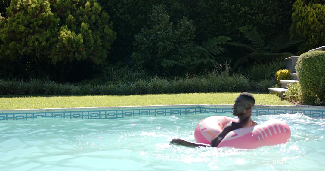 African American man floating on pink donut pool float in sunny backyard garden with copy space