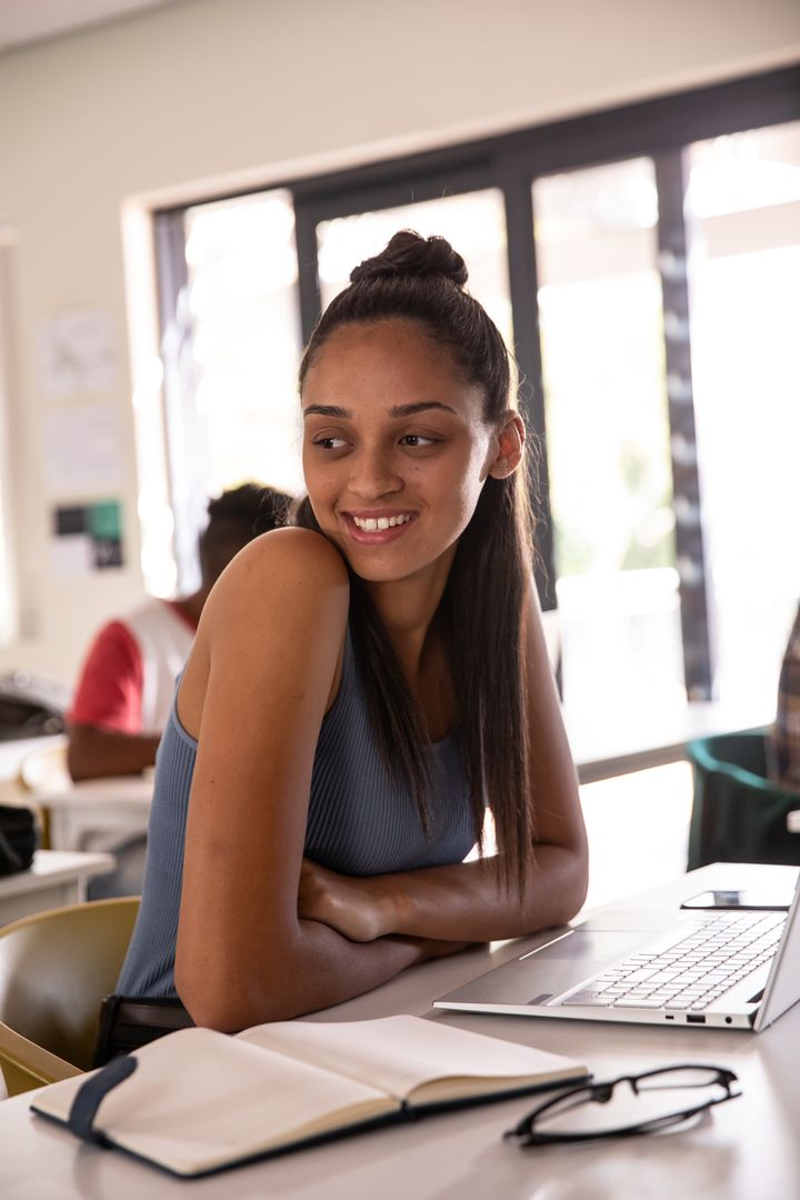 Smiling teenage student leaning at desk with laptop and open notebook in bright classroom