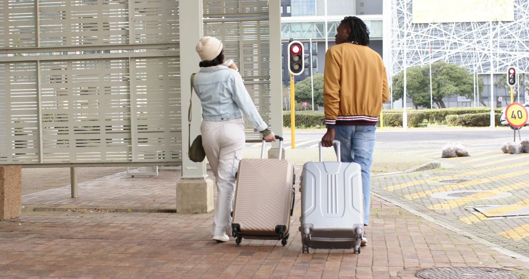 Diverse couple walking and pulling rolling suitcases at urban curbside shelter preparing for trip