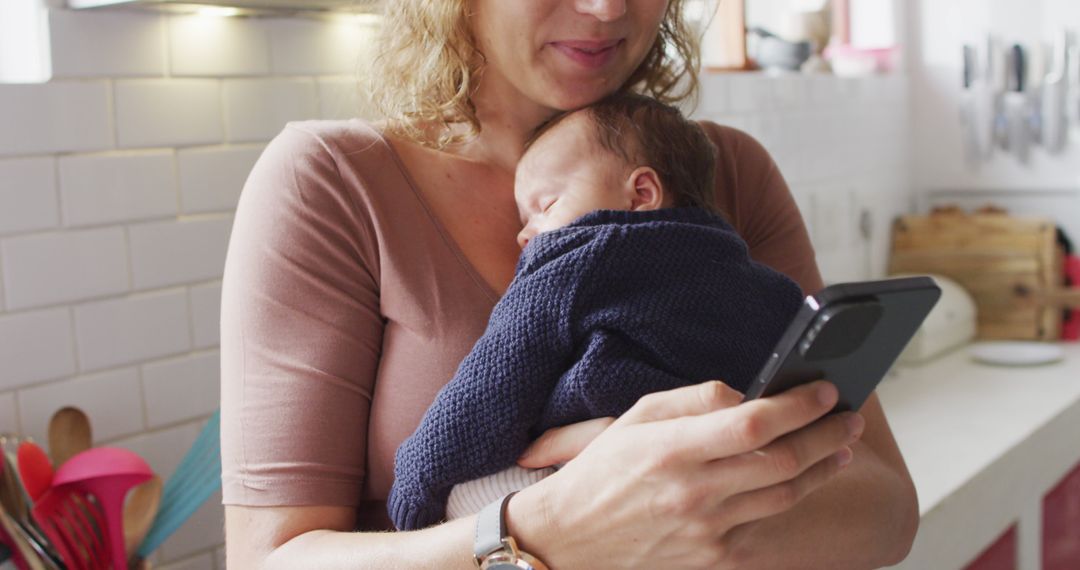 Mother holding newborn while using smartphone in cozy kitchen - Free Images, Stock Photos and Pictures on Pikwizard.com