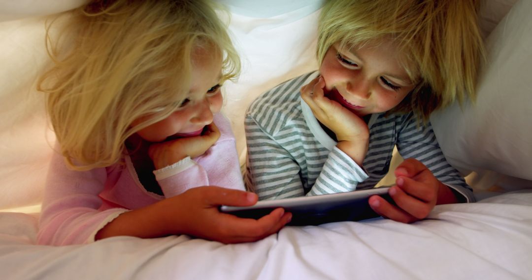 Happy Siblings Using Tablet Under Blanket Relax in Cozy Bedroom