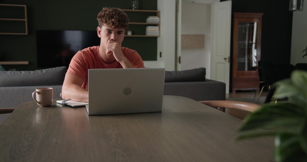 Young man working from home at laptop on dining table with coffee and notebook