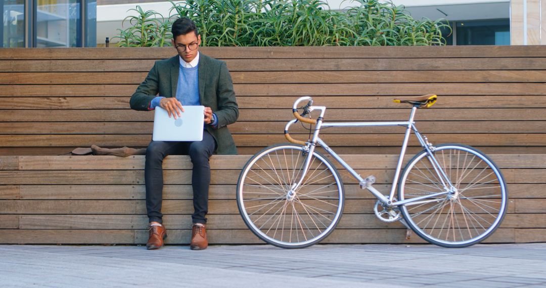 Man Working on Laptop While Sitting Outside on Wooden Bench with Bicycle - Free Images, Stock Photos and Pictures on Pikwizard.com