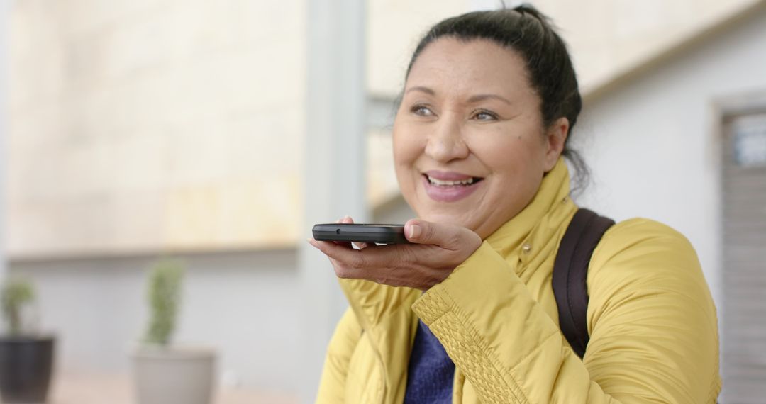 Smiling woman in yellow jacket using phone voice command while walking urban street