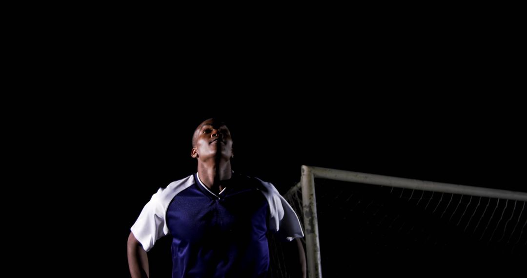 Soccer player gazing under stadium lights at night near goalpost and net