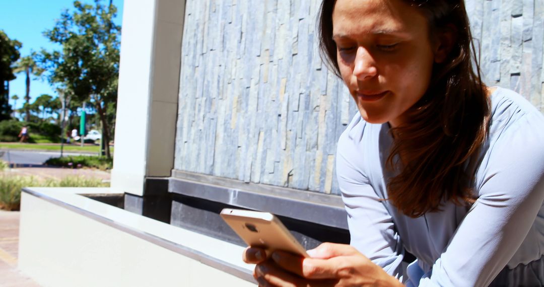 Woman Sitting Outdoors Using Smartphone Near Stone Wall on Sunny Day - Free Images, Stock Photos and Pictures on Pikwizard.com