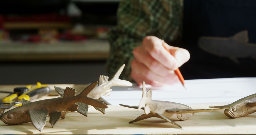 A middle-aged person is sketching on paper, surrounded by wooden fish sculptures, with copy space - Free Images, Stock Photos and Pictures on Pikwizard.com